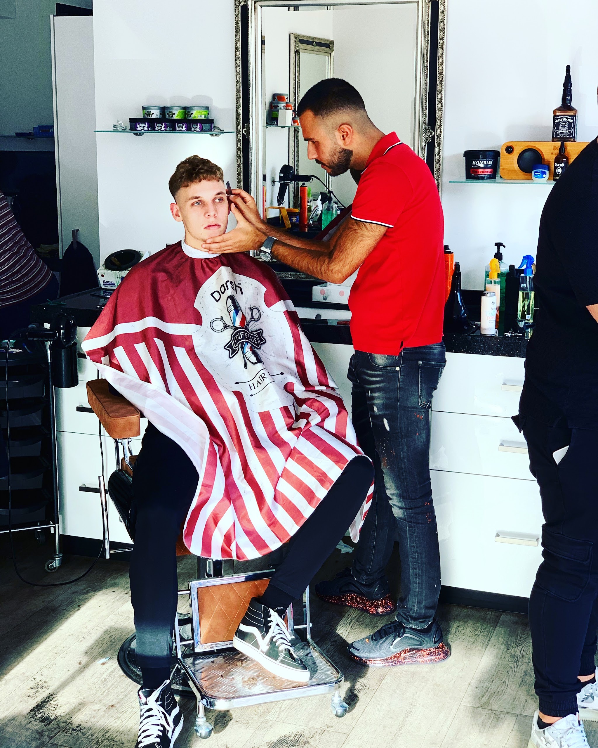 Kapper in een rood shirt werkt aan een klant in de stoel, onder een rood-wit gestreepte kapmantel