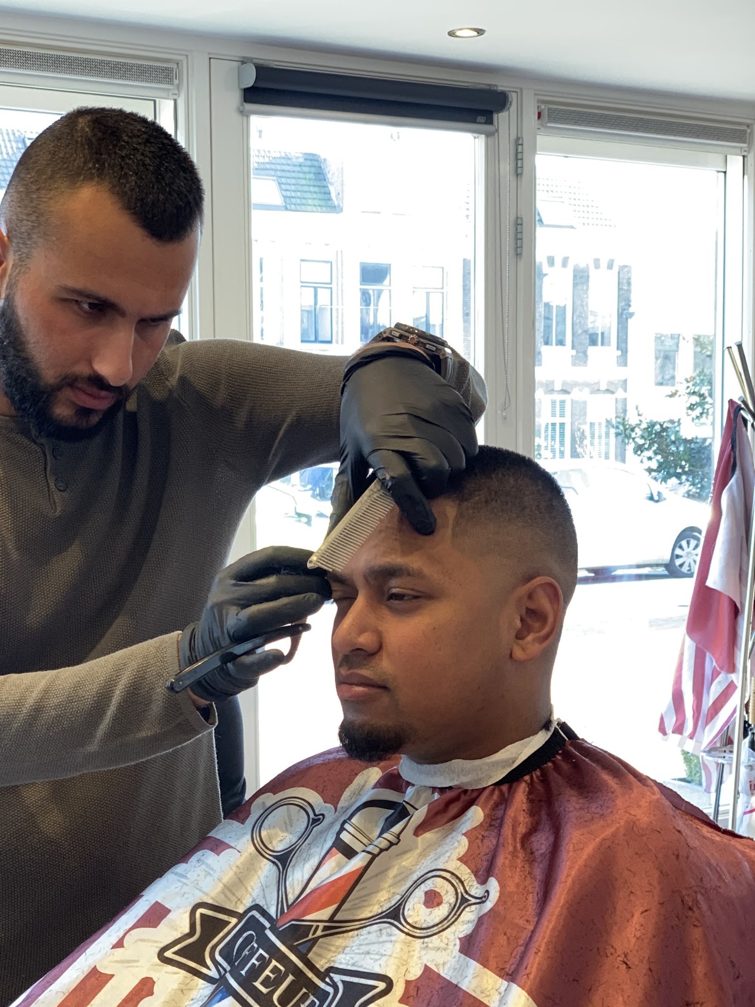 Kapper met zwarte handschoenen werkt met kam en tondeuse aan de haarlijn van een klant, gestreepte kapmantel