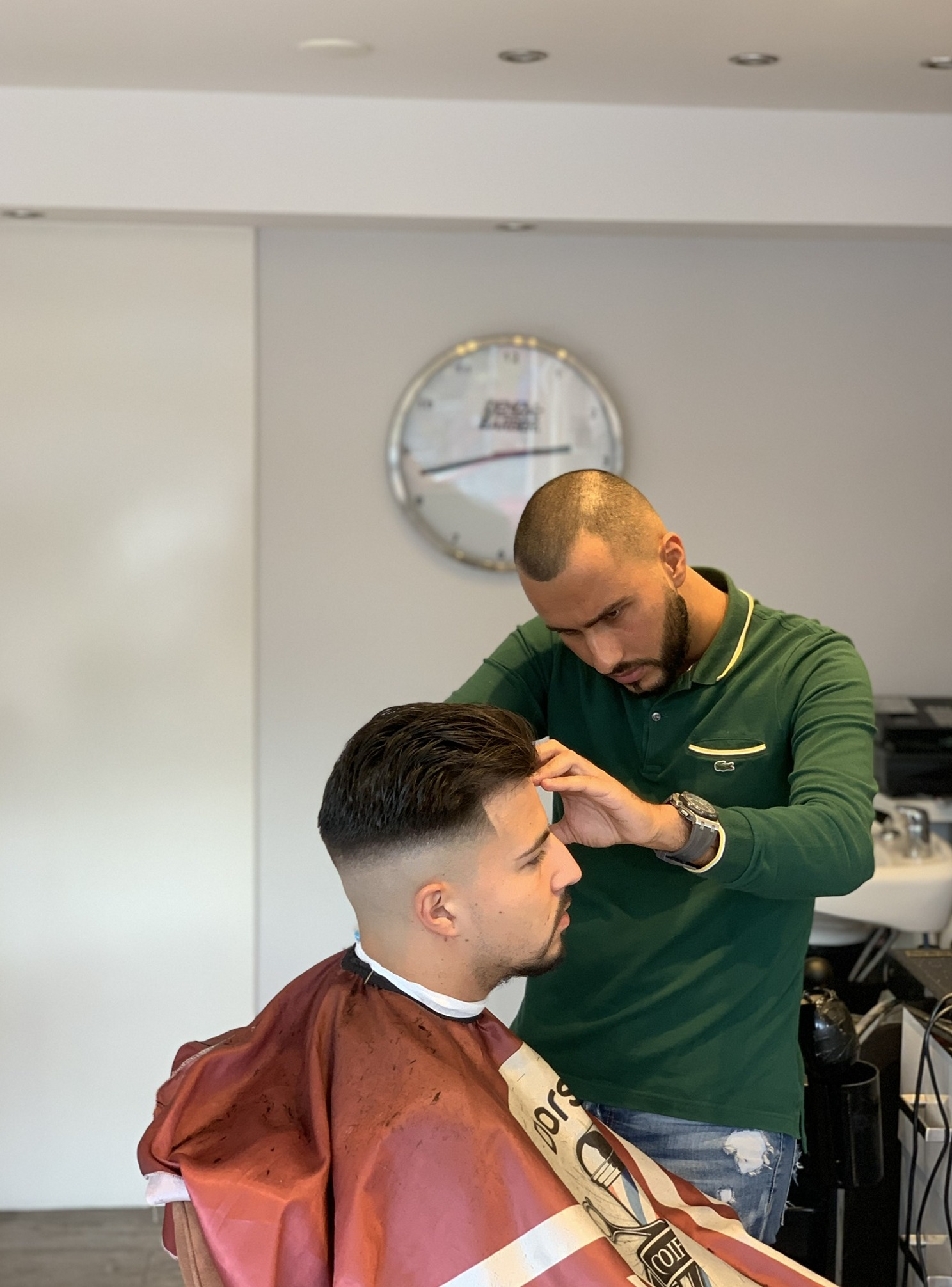 Kapper in een groen shirt maakt de bovenkant van een fade af, met de ronde wandklok erboven